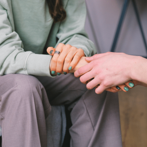 A woman wearing a light green sweatshirt and grey slacks having her hand held by a friend during a recovery resources meeting